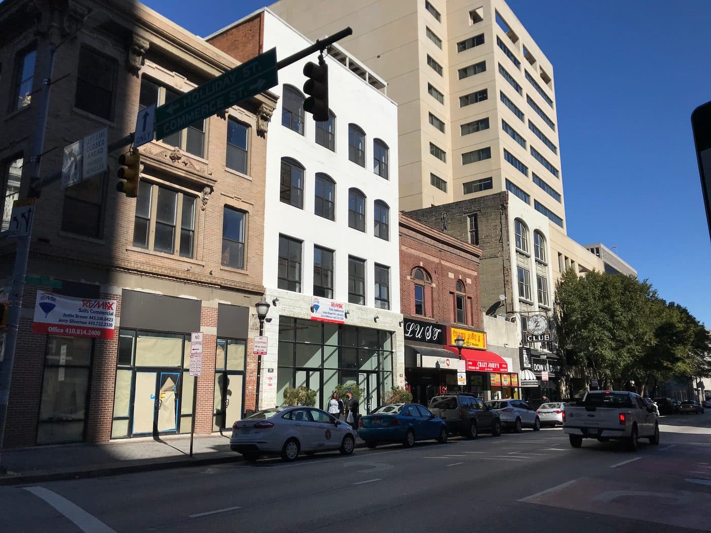 The 400 block of East Baltimore Street in 2019, with signs for Lust, Crazy John's and the 2 O'Clock Club along the right-hand side, and two empty shopfronts on the left papered over with commercial letting boards.