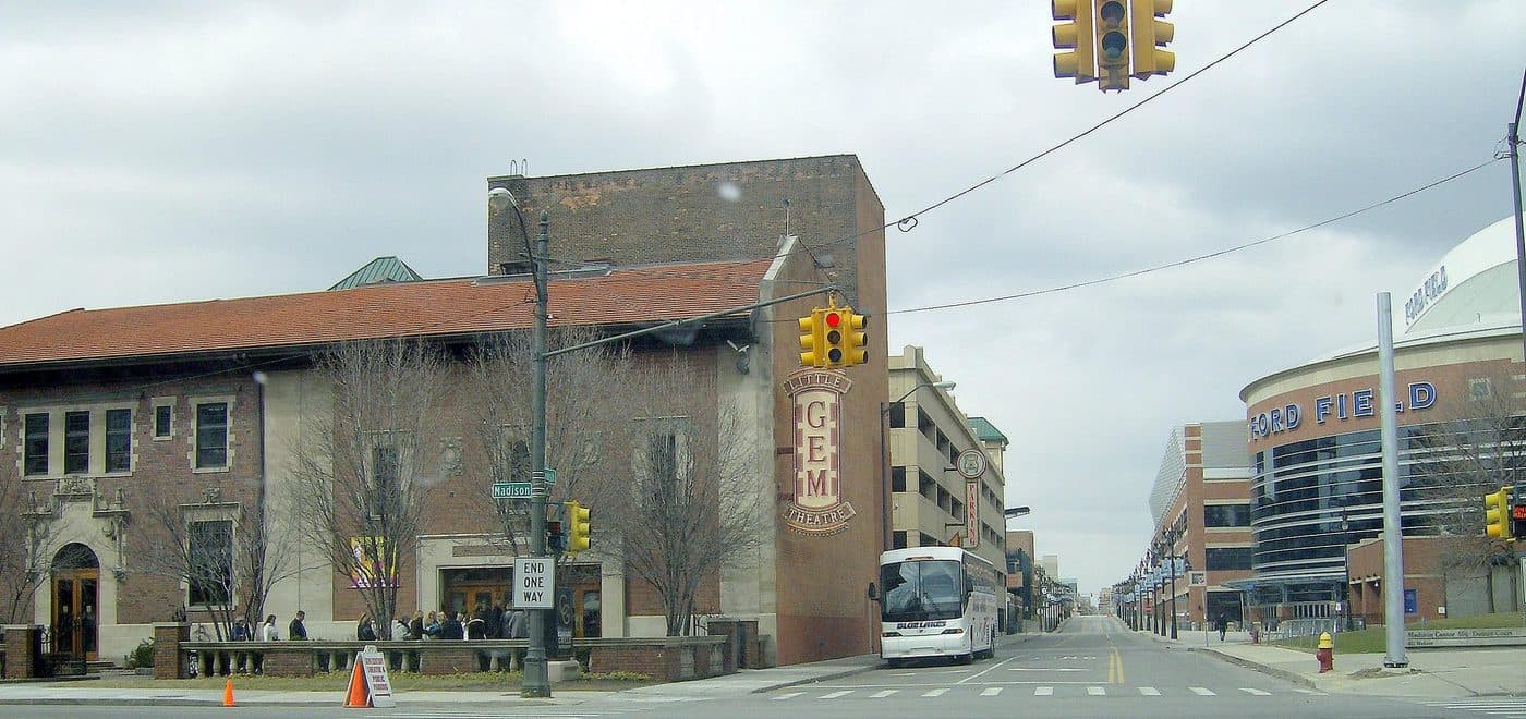 The Gem Theatre in Detroit at its current location on Madison Street, a low brick and terracotta building with a vertical sign reading Little Gem Theatre, with Ford Field visible down the street behind it.