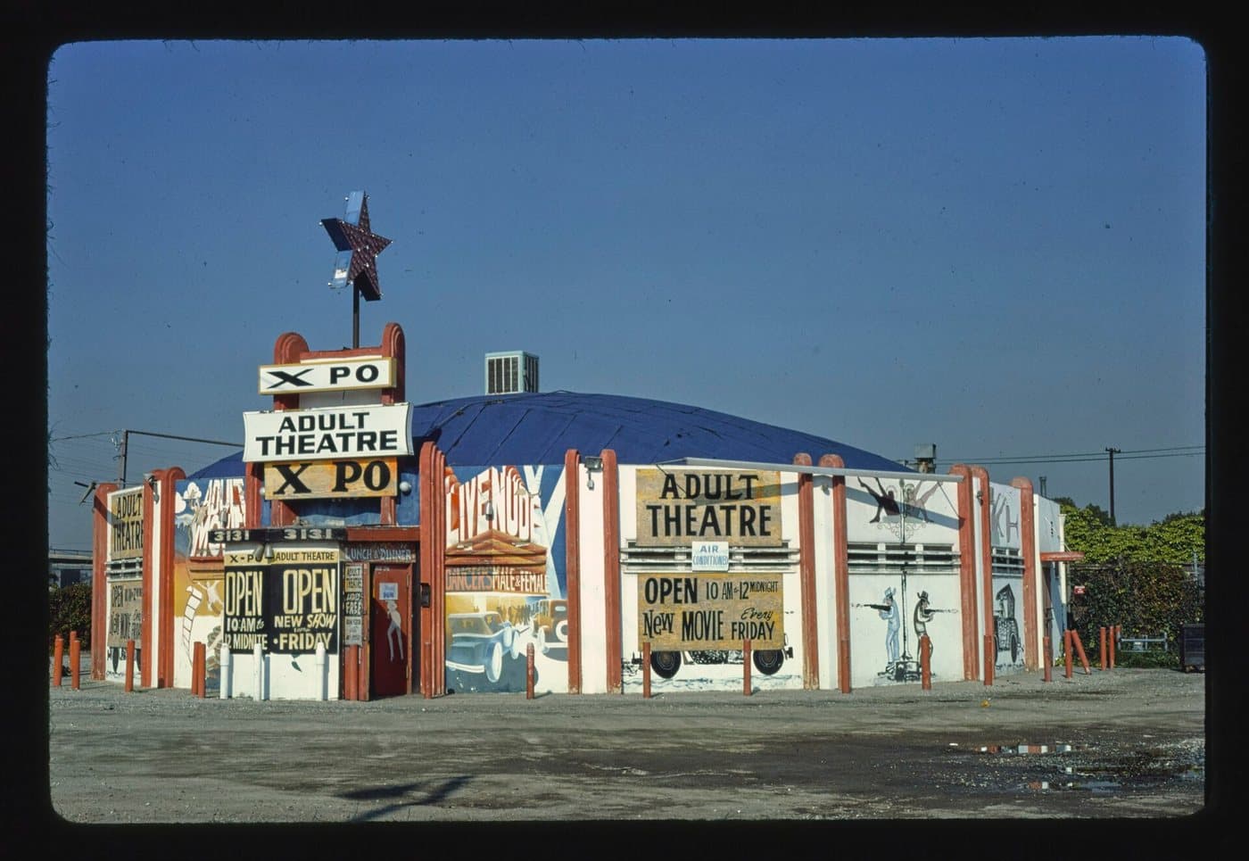 The XPO Adult Theatre in Wilmington, California, a low circus-tent-shaped building painted with murals, hand-lettered signs reading Adult Theatre, Open 10am to 12 midnite, and New Movie Every Friday, under a clear blue sky.