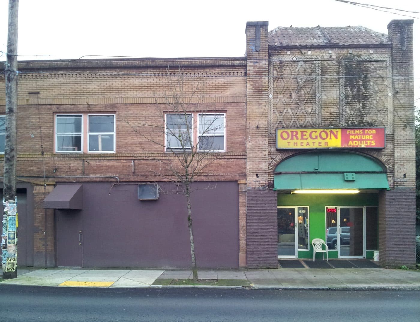 The Oregon Theater on Southeast Division Street in Portland in 2014, a brick building with a yellow and red sign reading Oregon Theater, Films For Mature Adults, and a lit neon open sign in the doorway.