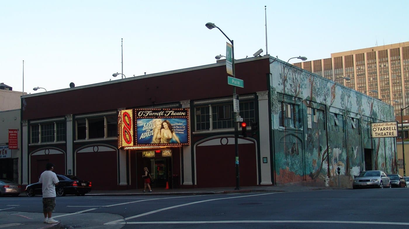 The Mitchell Brothers O'Farrell Theatre at the corner of O'Farrell and Polk Streets in San Francisco, its lit marquee advertising live shows, with a large painted mural covering the side wall.
